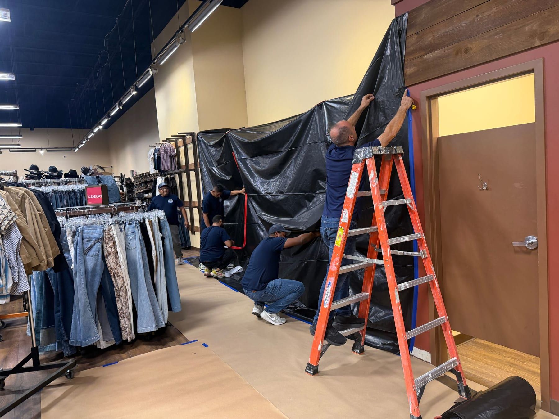 A floor-to-ceiling black plastic containment wall inside a store, two technicians on ladders securing the top edge, clothing racks behind them.