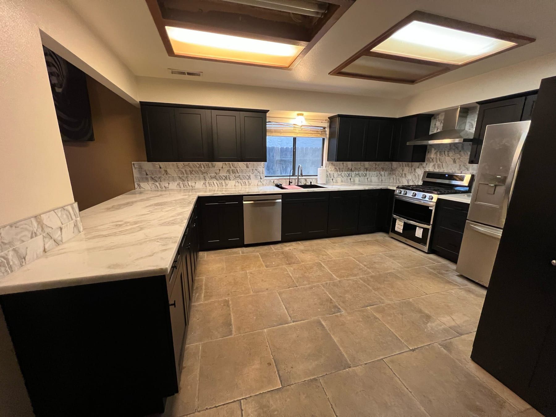 A U-shaped kitchen with dark cabinetry, marble-look countertops and backsplash, stainless appliances and a skylight above.
