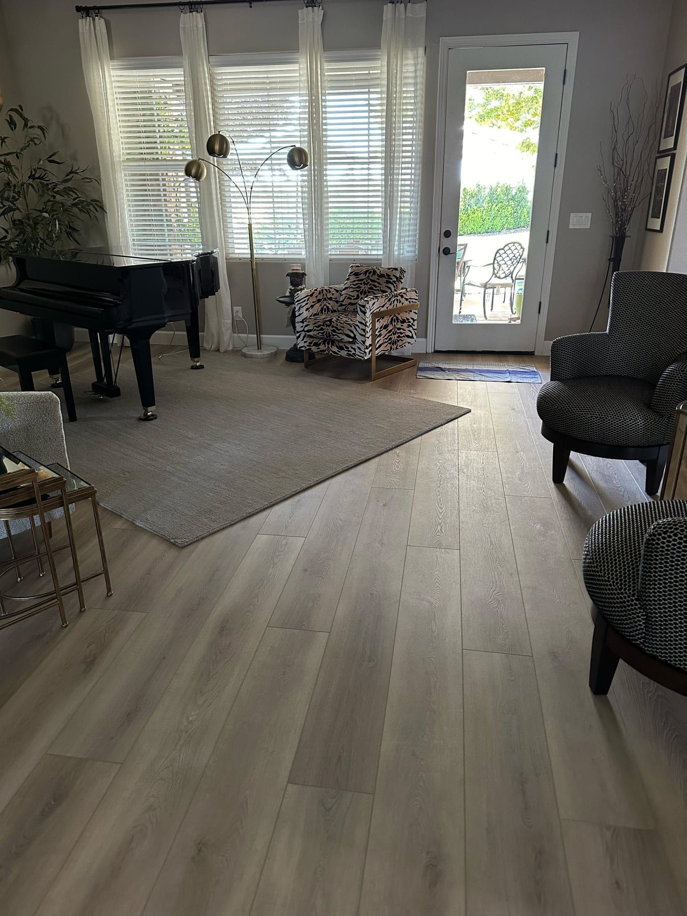 A living room with new light hardwood flooring, an area rug, a grand piano, and patterned armchairs near a window.