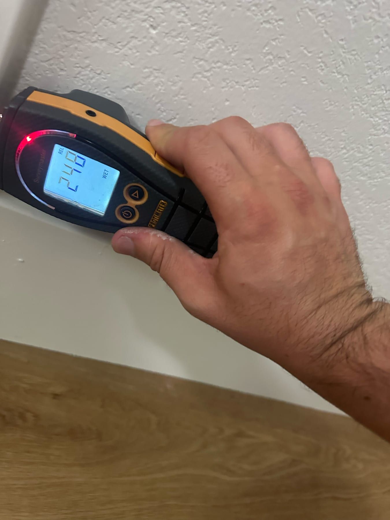 A hand pressing a moisture meter against an upper wall corner near the ceiling, red indicator lit and display marked wet.
