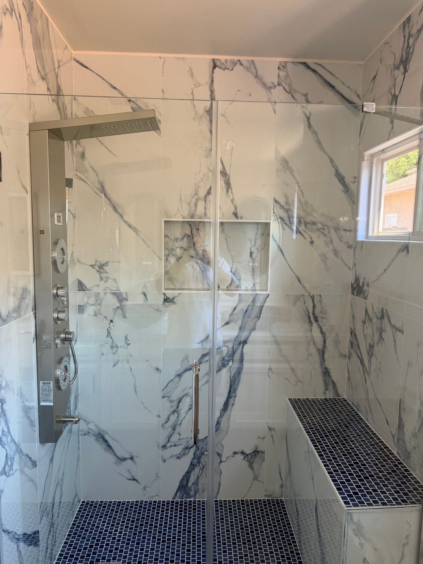 A marble-tiled walk-in shower with a blue glass mosaic bench, a stainless shower panel, and a glass door.