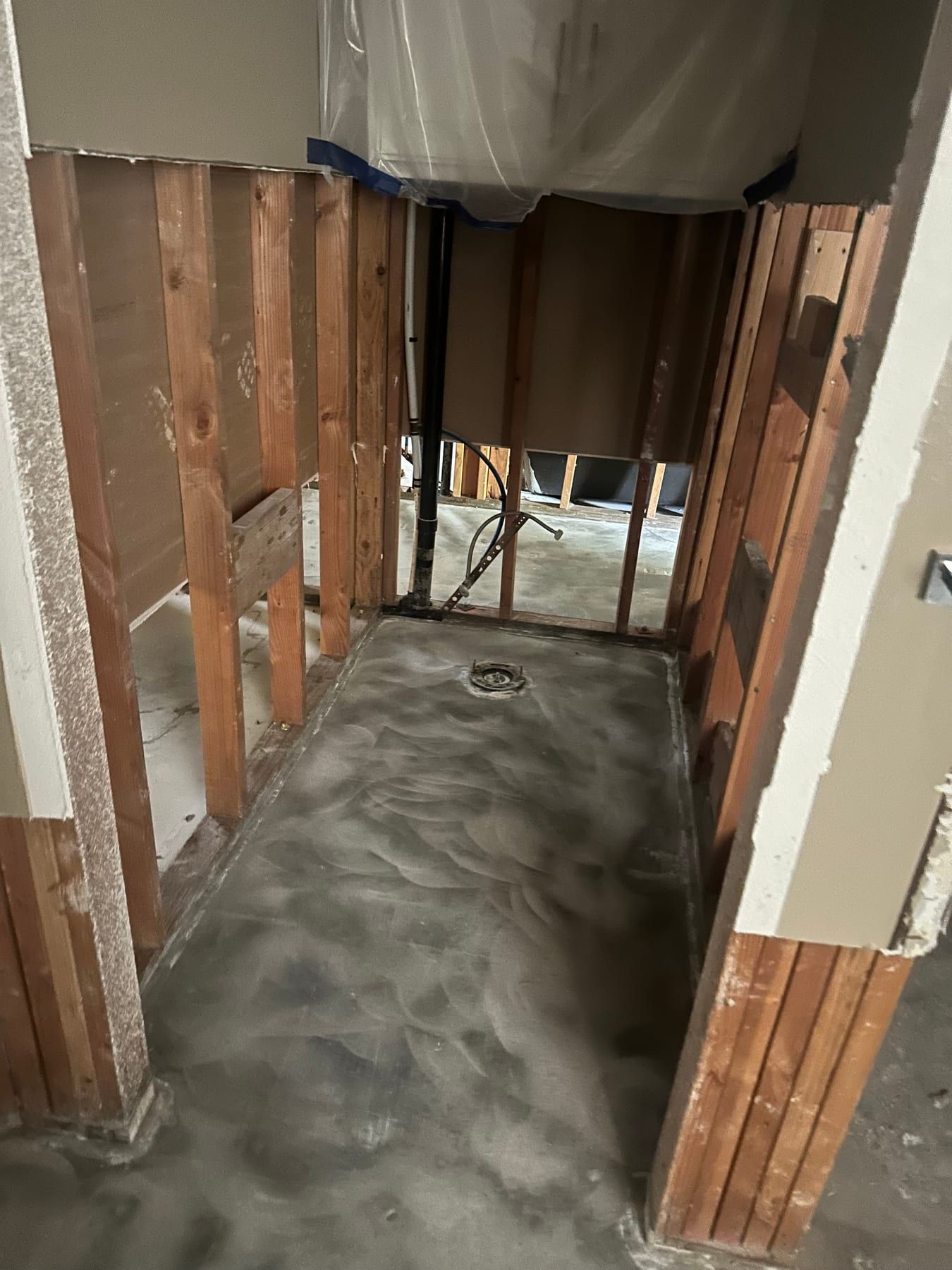 A narrow shower stall stripped to bare wood studs and concrete, drain exposed, plastic sheeting draped over the ceiling opening.