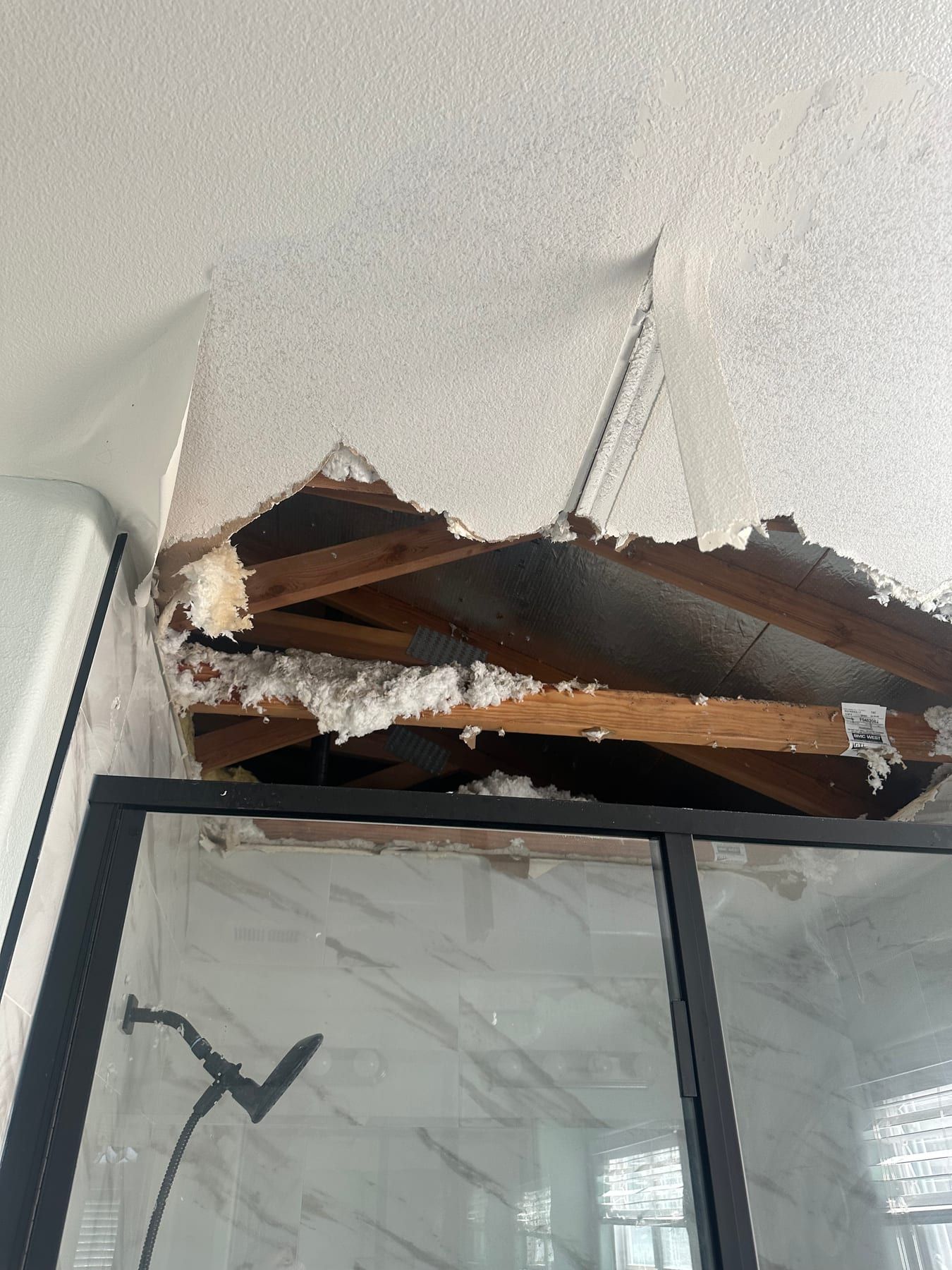 A hole in a textured ceiling above a glass shower enclosure, with roof framing and loose insulation hanging through the opening.