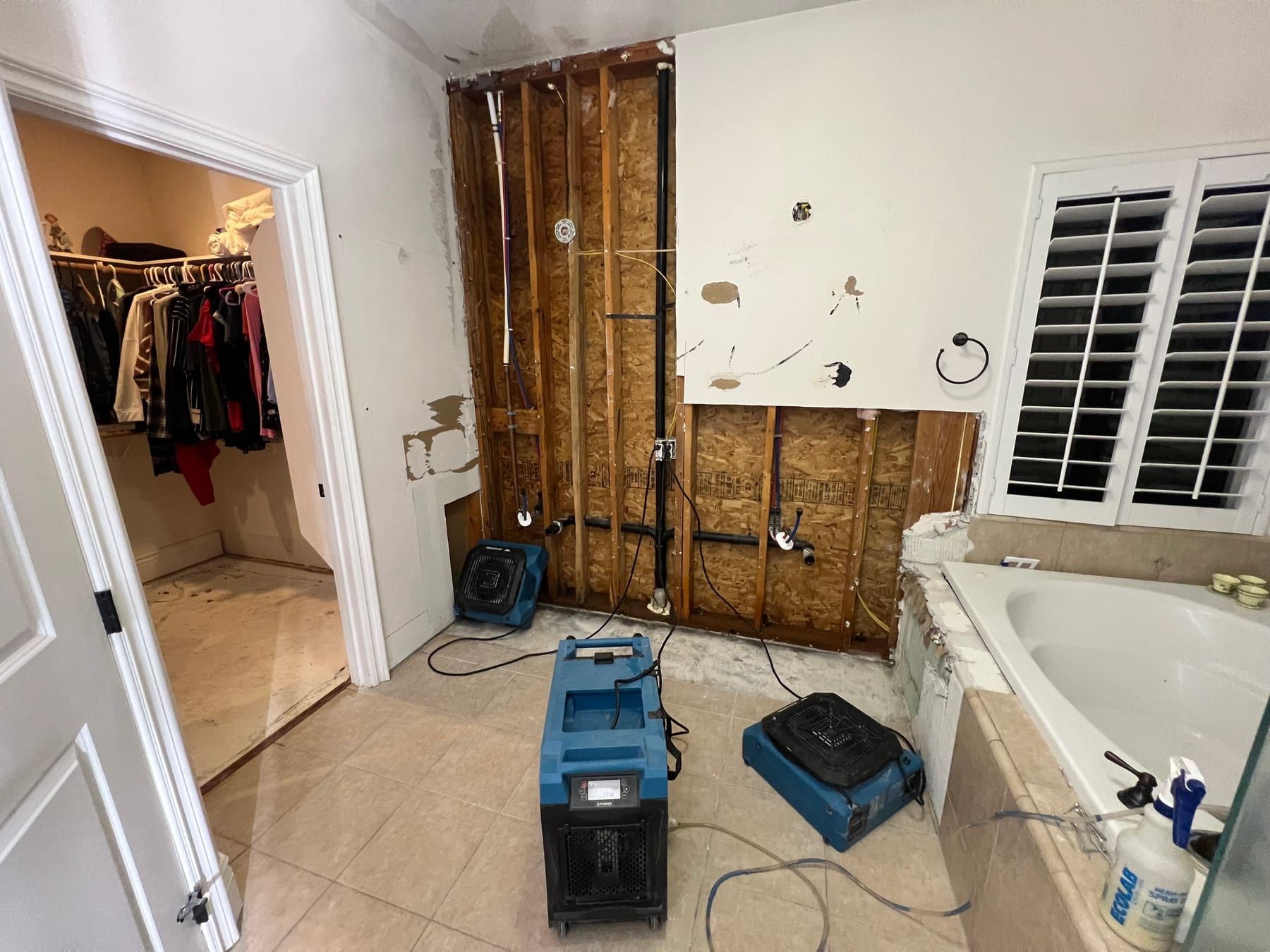A blue dehumidifier and two air movers running on a tile bathroom floor in front of a wall opened to the studs, bathtub to the right.
